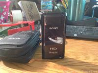 Front view of the purple Sony HD bloggie video camera and black carrying case on a wooden table.