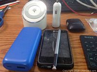 Photo showing various electronic items including a white Qi wireless charger, white portable speaker, Samsung battery, blue power bank, LG smartphone, and a stylus pen.