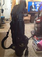 Side view of the black Masters golf bag with shoulder strap and embroidered logo, some golf clubs visible inside with black head covers and parts of a room around it