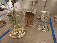 Three mantel clocks on a flat surface including two anniversary clocks with glass domes and one rectangular clock.