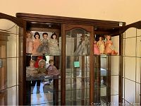 Full display cabinet showing 11 Madame Alexander dolls arranged in two rows, several in pink and silver dresses.