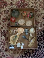 Full view of glass and stone paperweights and carved stone figures arranged in two cardboard boxes on a patterned rug.