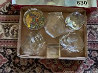 Top view of five multifaceted clear glass paperweights, one small clear cube paperweight, and a round paperweight with butterfly and floral design inside.