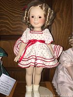 Front view of Shirley Temple porcelain doll 14 inches tall in white dress with red polka dots and red ribbon trim and curly blonde hair with red bows.