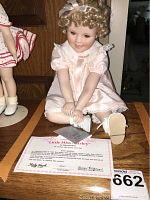 Front view of Little Miss Shirley porcelain doll seated on wooden surface, showing pink dress, curly hair, white shoes, certificate and tag.