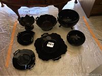 Black glassware assortment of seven pieces shown arranged on protective floor covering beneath furniture. Pieces include bowls and plates with decorative scalloped edges and handles.