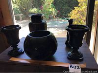 Photo showing five black art glass items on a dark surface with garden background. Includes bowl, vases, and goblets.