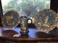 Photo showing two collectible plates and one stein displayed on a wooden surface by a window with natural daylight. Plates have detailed Snow White and Seven Dwarfs scenes on blue background with floral and gold designs, stein shows similar design with pewter lid.
