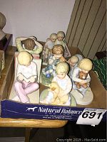 Nine Francis Hook figurines of children in various poses inside a Natural Balance dog food box on a wooden table.