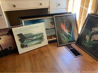 Collection of 3 unframed oil paintings and 3 framed floral prints arranged on wooden floor near drawers