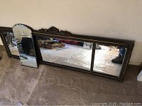 Large rectangular mirror with ornate carved frame and smaller scalloped mirror leaning against a wall on carpeted floor