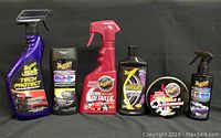 All six Meguiar's car care products lined up side by side on a dark surface with black background, showing product labels and bottle shapes.