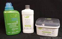 Image of three Amway Legacy of Clean laundry products side by side on black background: Fabric Softener 33.8oz (green bottle), Prewash Liquid 16.9oz (white bottle), and All Fabric Bleach 2.6lbs (white tub with gray lid).