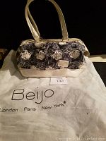 Full front view of BEIJO embroidered handbag with white floral and leaf design, white leather-like bottom and handles, placed on white branded carrying bag.