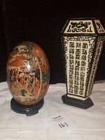 Photo of the decorative Chinese egg sculpture and hexagonal Chinese vase side by side on black stands against a black background.