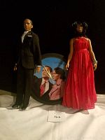 Two porcelain dolls and a wall clock with Barack and Michelle Obama photo face on a dark background.
