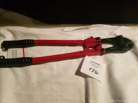 Full view of the red-handled bolt cutter with black grips and black cutting head lying on a white cloth with a MaxSold tag.