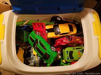 View looking down into the open blue and yellow plastic tote showing a collection of toy cars and trucks in assorted bright colors including green, red, yellow, and black.