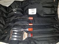 Photo showing three BBQ utensils (spatula, fork, tongs) with wooden handles lying in a black fabric pouch.
