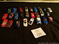 Full view of all approximately 26 Hot Wheels cars laid out on black cloth, showing a variety of colors and styles including race cars, classic cars, and trucks.