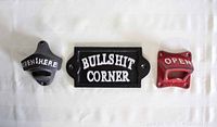 Three cast iron items arranged on a white surface: two wall mount bottle openers in red and black colors, and one black sign with white text 'BULLSHIT CORNER'.