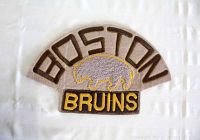 Top down view of Boston Bruins embroidered crest patch featuring the 1928 style bear logo and text 'BOSTON' and 'BRUINS' on grey fabric backing with gold and brown stitching.
