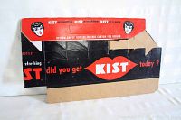 Photo of front side of flat vintage Kist cardboard bottle carrier showing red, black and white logo and face graphics with text.