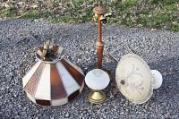 Four vintage light fixtures outside on gravel including a tall wooden table lamp stand, a small table lamp with frosted white glass shade and brass base, and two ceiling lights including a hanging stained glass fixture.