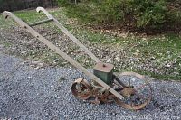 Side view of antique manual seed planter showing wooden handles, green metal seed hopper, spoked front wheel and rear wheel with seed dispensing mechanism.