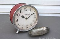 Front view of red rustic-style clock with white dial, black numbers, and silver edges, with small vintage silver dish beside it