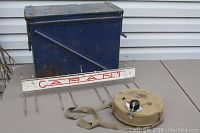 View of blue metal ammo box, white Carant metal hook rack, and camping water canteen on floor in front of white outdoor siding