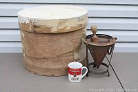 Full view of the wooden cheesebox with lid, metal kitchen strainer with wooden press, and Campbell's Soup mug placed on the ground.