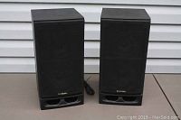 Pair of black Samsung speakers side by side showing front with fabric grille and bass reflex port.