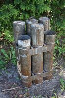 Front view of six attached tall metal canisters with visible rust and wear, standing upright outdoors on dirt ground.