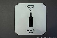 Front view of square white metal sign showing black wine bottle silhouette with Wi-Fi symbol above and 'Wine Fi Zone' text below.