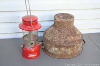 Red Coleman lantern and a rusty metal milk can funnel/strainer side by side on a surface in front of a house wall.
