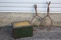 Photo shows a green metal ammo box with a hinged lid and latch on the front, sitting on gravel in front of a white wall. To the right are two large rustic iron ice tongs with scissor handles and claw-like gripping ends.