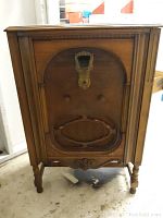 Front view of antique wooden floor-standing radio cabinet with carved wood details and damaged front grill.