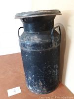 Full view of black metal milk can with signs of wear and rust on the lid and body.