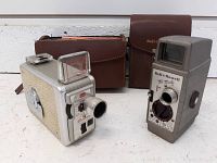 Both Kodak and Bell & Howell movie cameras side by side with their brown leather carrying cases behind them.