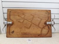 Front view of wooden beef cutting board with longhorn metal handles. The surface shows a carved design and juice groove edges.