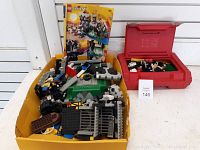 Yellow tray with assorted Lego castle pieces including minifigures, parts of walls, and flags. Instruction manual visible at back. Red plastic carry case to the side containing additional Lego pieces.