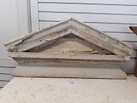 Front view of triangular wooden exterior window cornice with peeling white paint on an indoor surface against a white wall.