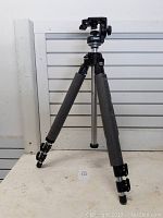 Full view of black Manfrotto tripod with foam grips on two of the legs, set on a table in front of a white slat panel wall.