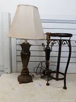 Two metal lamps and one metal plant stand shown side by side against white slat wall. One lamp has beige shade; other lamp does not. Plant stand is dark metal with decorative details.