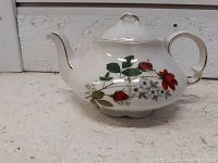 Porcelain teapot showing rose floral pattern on one side, curved spout, and gold trim on handle and lid.
