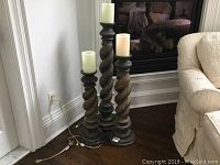 Three graduated electric candlesticks displayed side by side on a hardwood floor near a white wall and beige sofa.