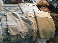 Photo showing three custom-made drapes laid out on a bed, two beige with grommets and one off-white with stitched border.