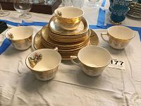 Photo of multiple tea cups, saucers, and plates stacked on a table showing the 22K gold trim and painted historical scenes on cups.