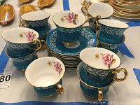 Eight teal blue porcelain cups with gold handles and pink rose decorations, atop a table with some saucers underneath